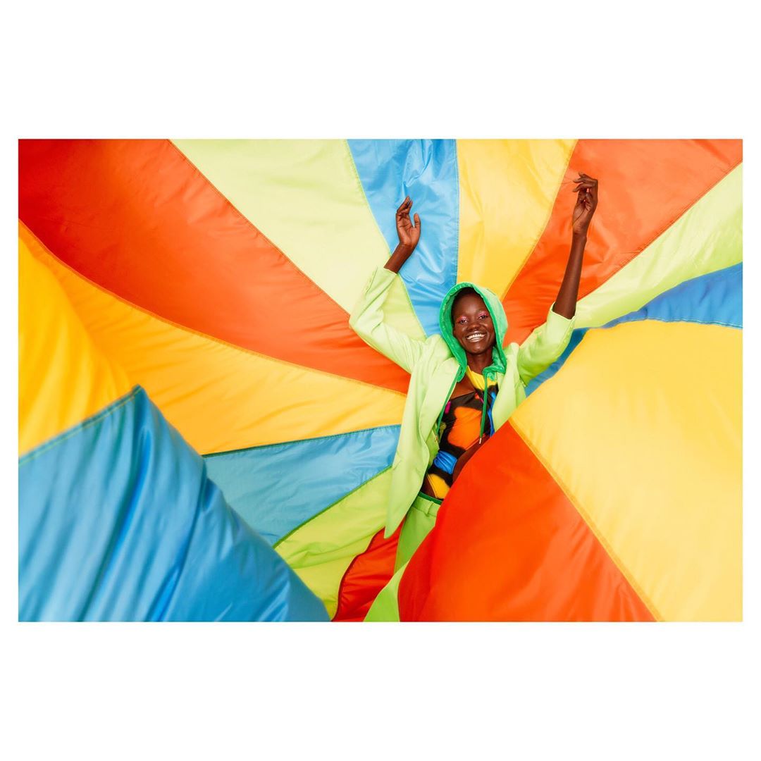Woman in neon green hooded tracksuit poses with arms raised against a vibrant backdrop of blue, orange, and yellow geometric shapes.