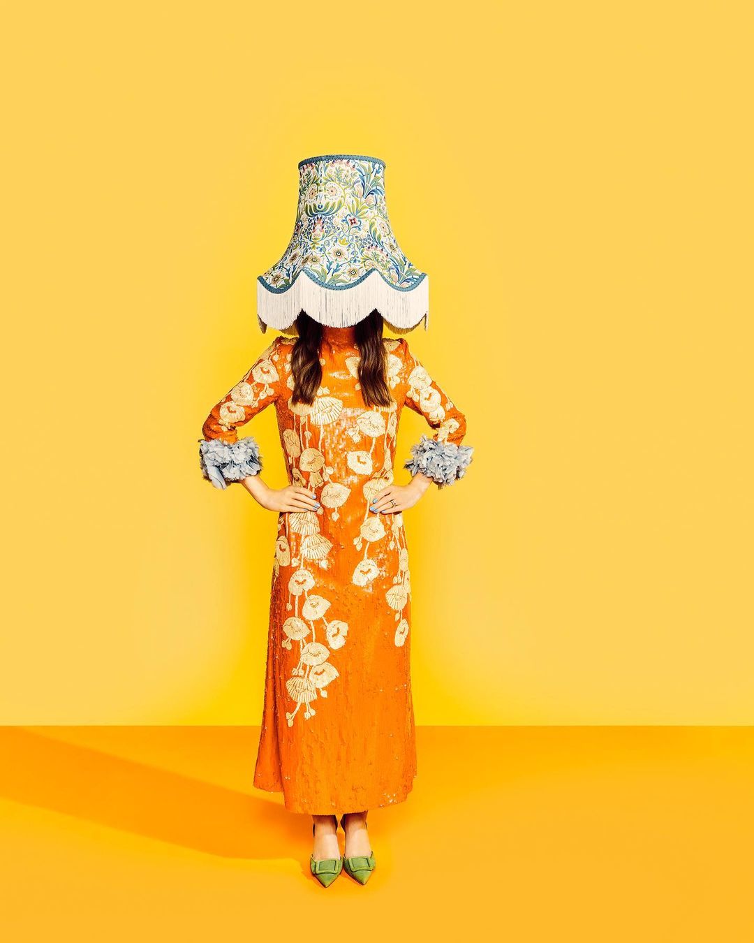 Woman in orange dress with floral details and a lampshade on her head against a yellow backdrop for a surreal fashion shot.