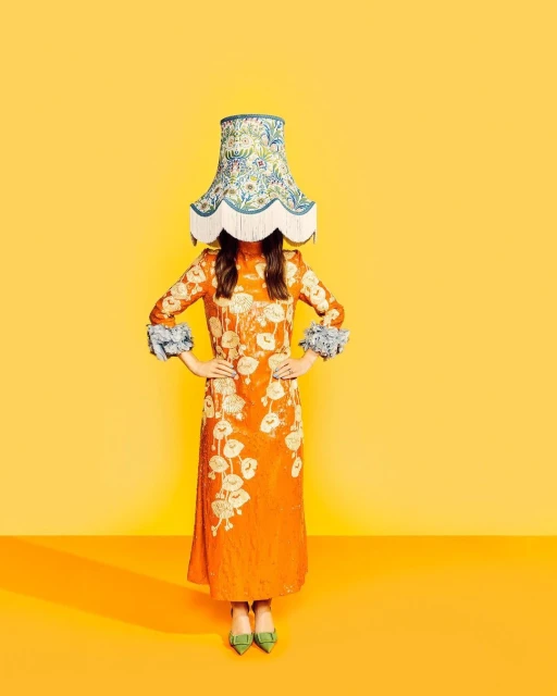 Woman in orange dress with floral details and a lampshade on her head against a yellow backdrop for a surreal fashion shot.