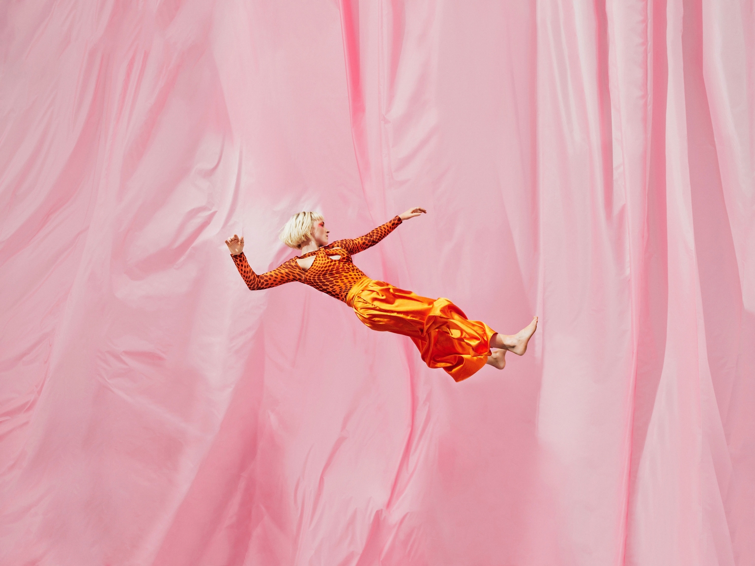 A woman with short blonde hair in a patterned long-sleeve shirt and orange pants is floating mid-air against a draped pink background.