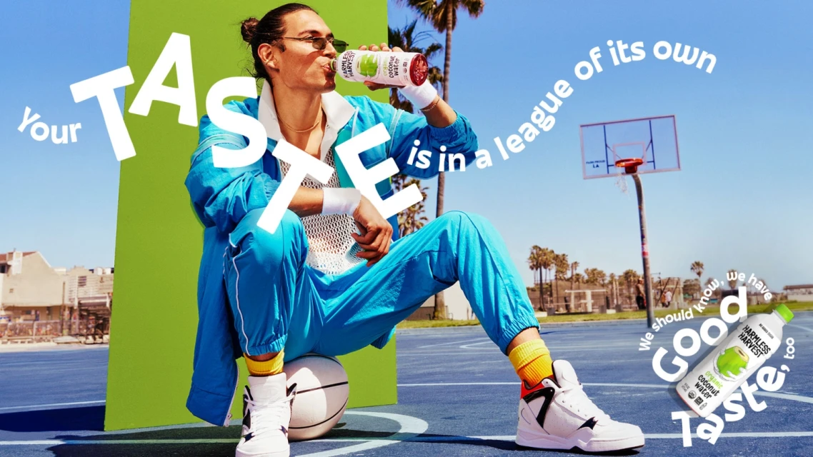 Man in a blue tracksuit sitting on a basketball, drinking Harmless Harvest coconut water, with a basketball hoop and palm trees in the background.