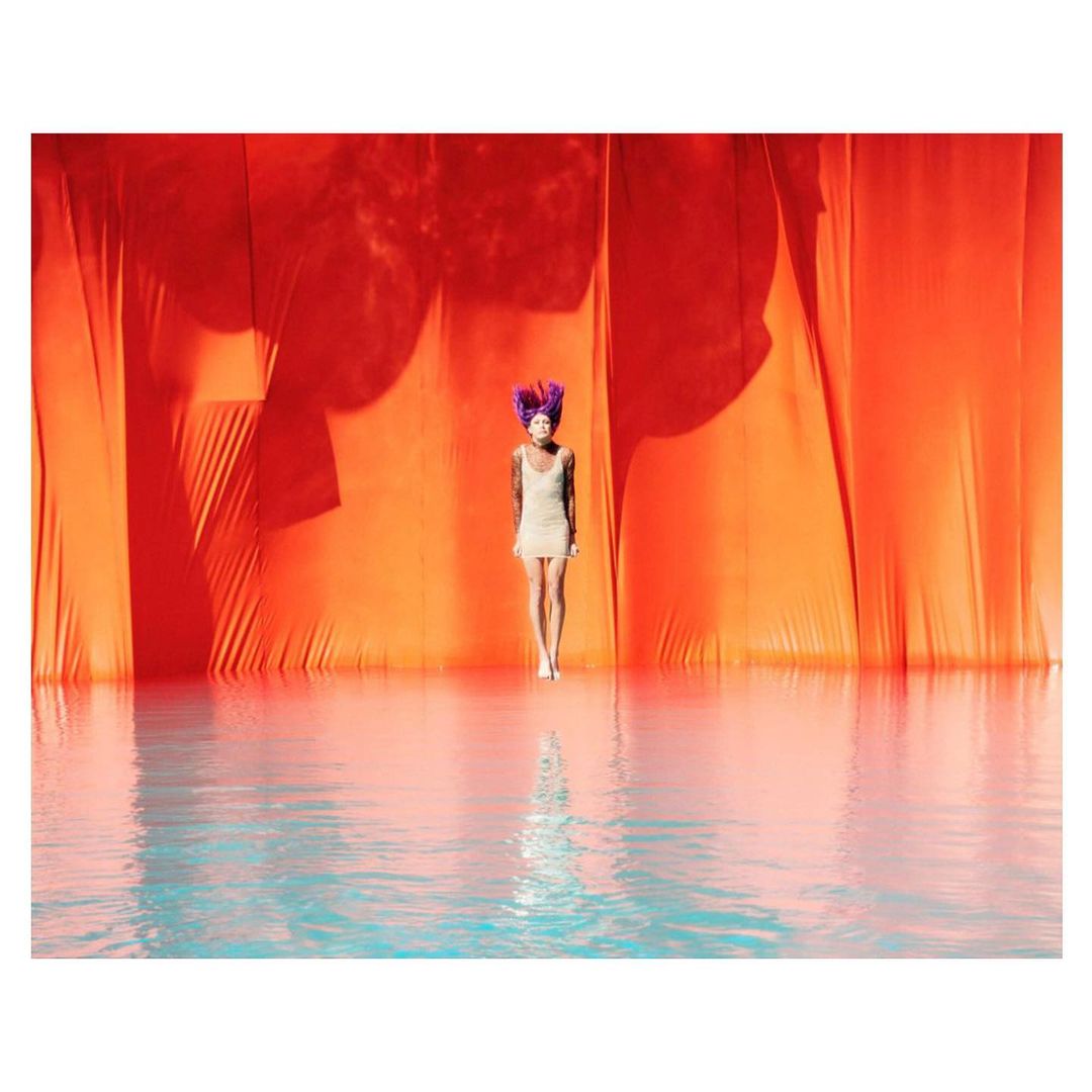 Woman with purple hair stands in front of an orange drape in a water-covered area, reflecting her image.