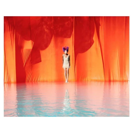Woman with purple hair stands in front of an orange drape in a water-covered area, reflecting her image.