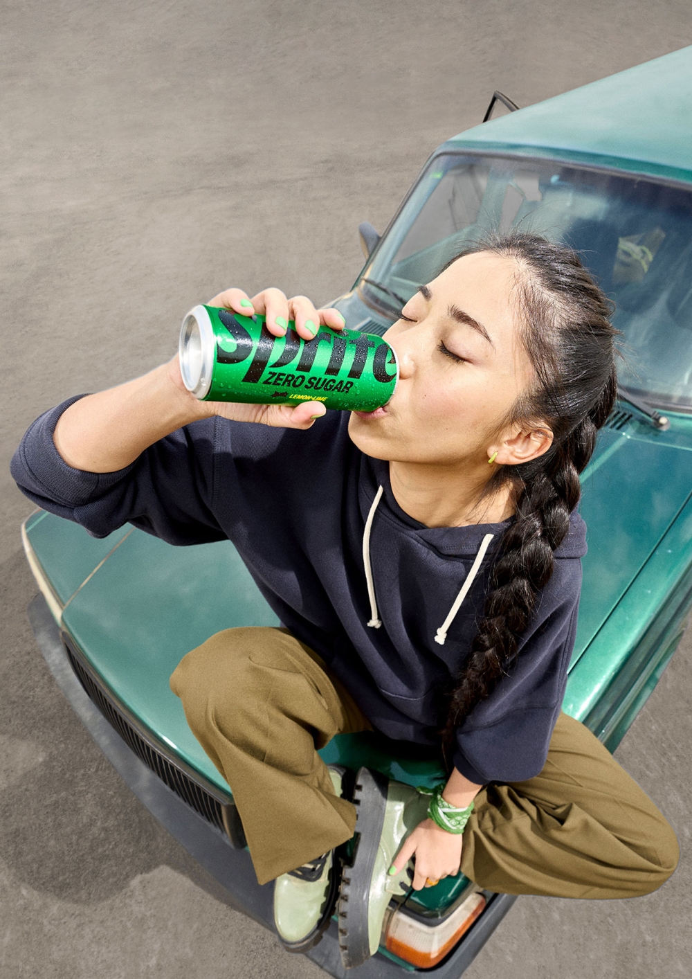 Young woman drinking Sprite Zero Sugar while sitting on the hood of a green car with concrete background, promoting drink brand.
