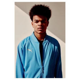 Young man with an afro hairstyle wearing a blue zip-up Puma tracksuit jacket against a neutral wall with diagonal shadows.
