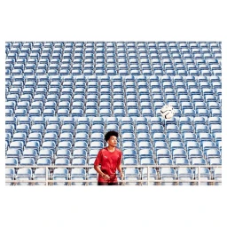 Young man in a red sports jersey standing in front of endless rows of empty blue stadium seats with a soccer ball floating in the air.
