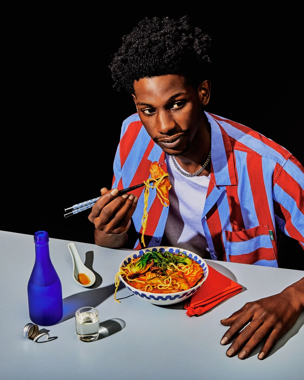 Young Black man in a striped shirt sits at a table, eating noodles with chopsticks. Bottle and glass of liquid and red napkin on table.