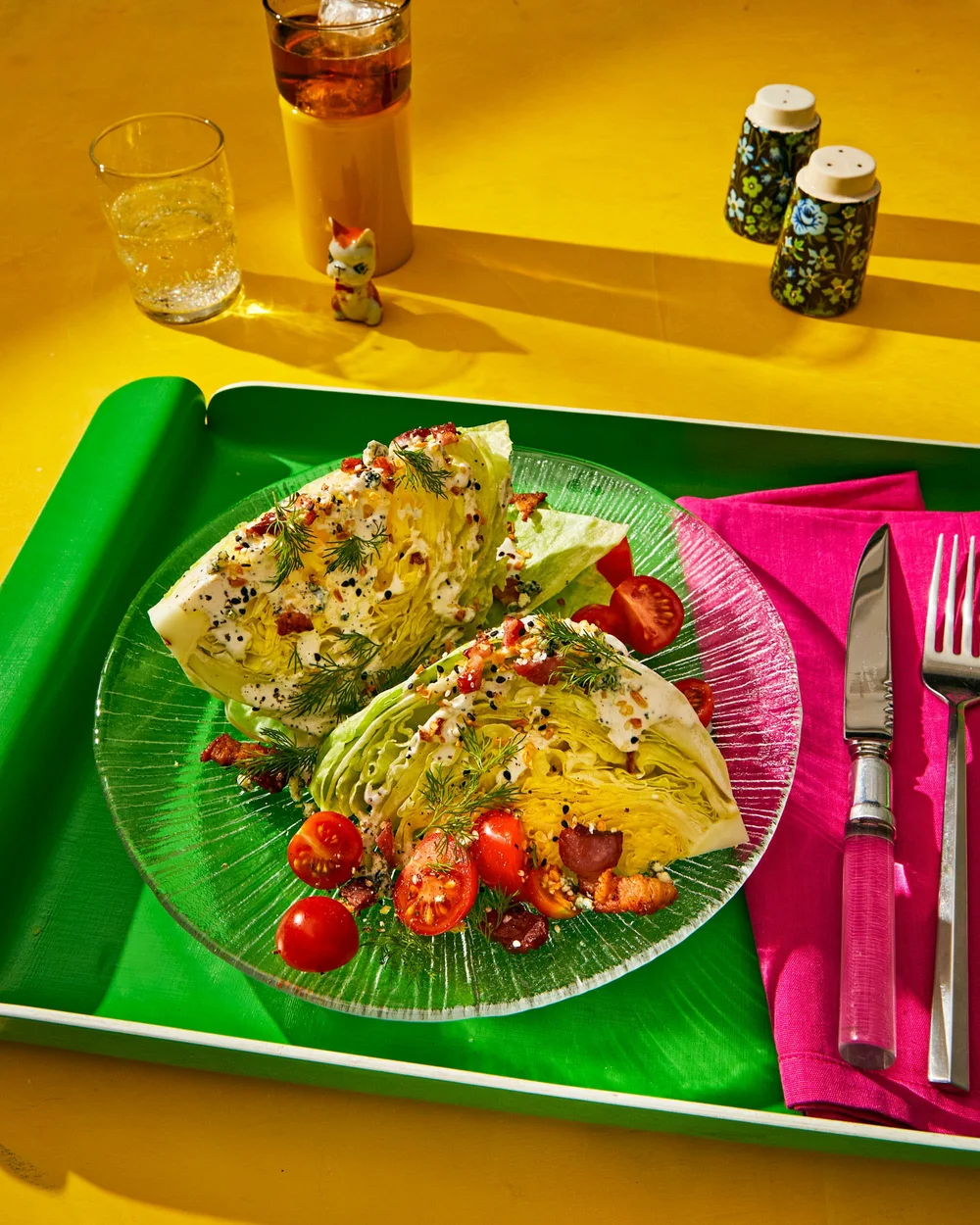 Overhead shot of wedge salads on a green tray with drinks, floral shakers, and pink napkins, on a yellow surface.