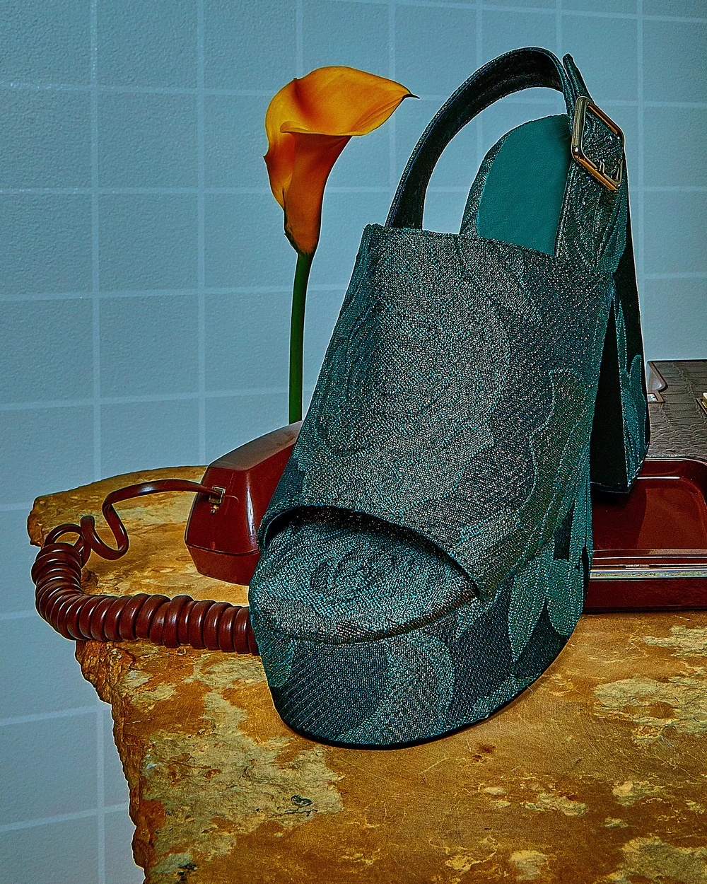 Stylish high-heeled platform sandal with a green floral pattern sits next to an orange calla lily and a vintage rotary phone on a stone surface.