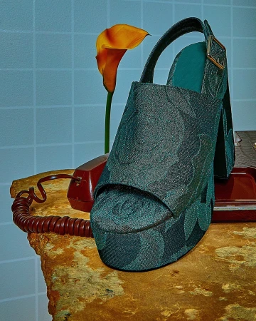 Stylish high-heeled platform sandal with a green floral pattern sits next to an orange calla lily and a vintage rotary phone on a stone surface.