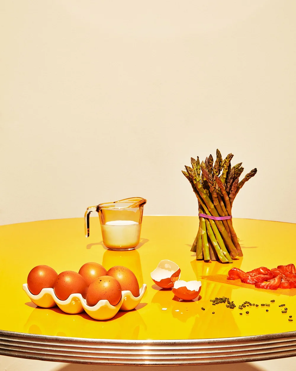Still life photo of eggs in a carton, asparagus bundle, measuring cup with liquid, eggshells, red food pieces on a yellow table.