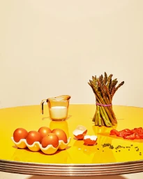 Food still life with an asparagus bundle, brown eggs in a holder, and a measuring cup of milk on a bright yellow table.