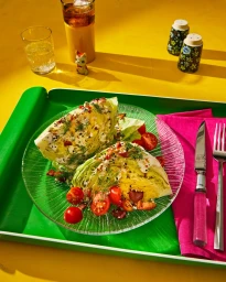 Two dressed iceberg wedge salad portions with cherry tomatoes on a textured glass plate, set on a green tray over a yellow surface.