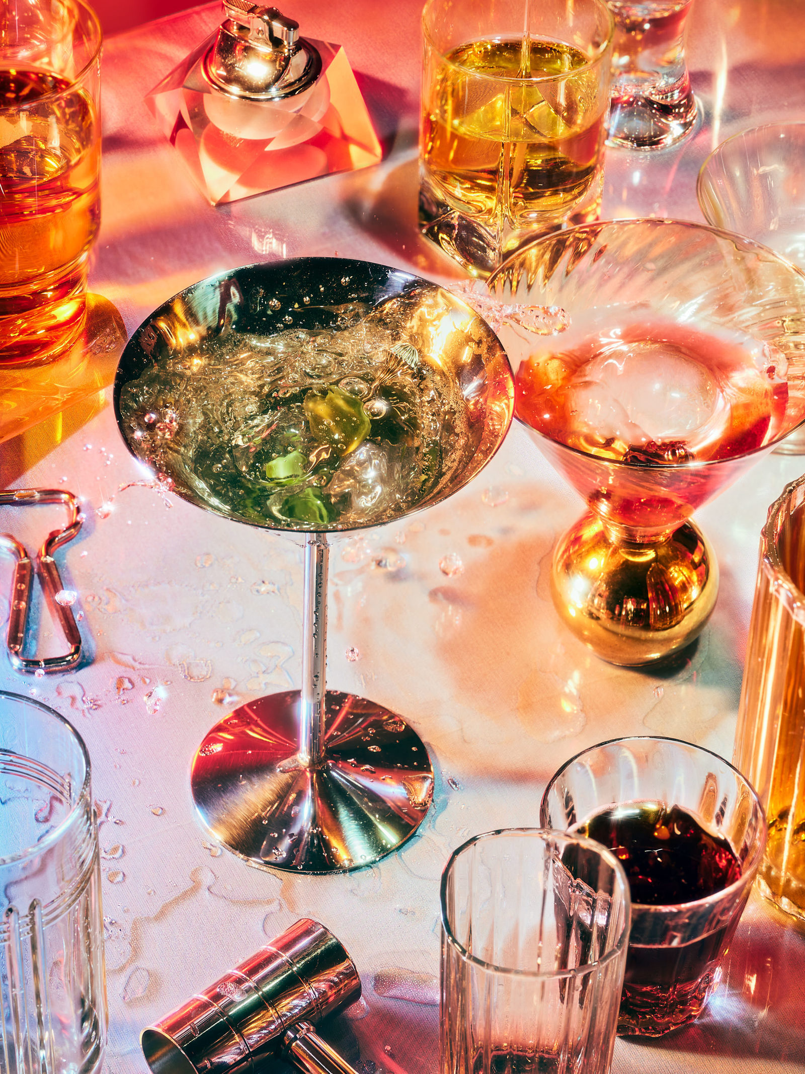 Various cocktail glasses filled with colourful drinks and bar tools are arranged on a reflective surface with a pink and blue lighting.