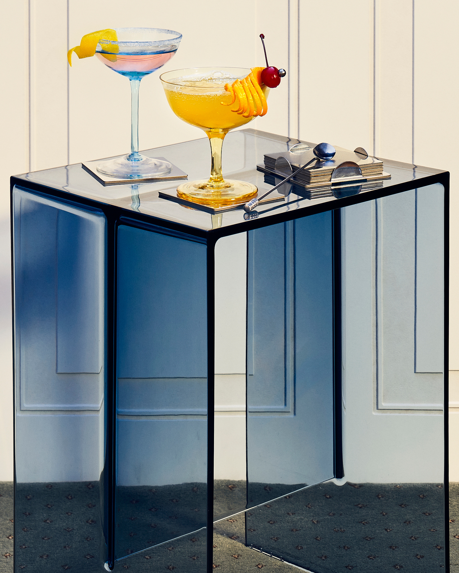 Two cocktail glasses with orange and cherry garnishes sit on a blue translucent table against a cream-colored wall with simple molding.