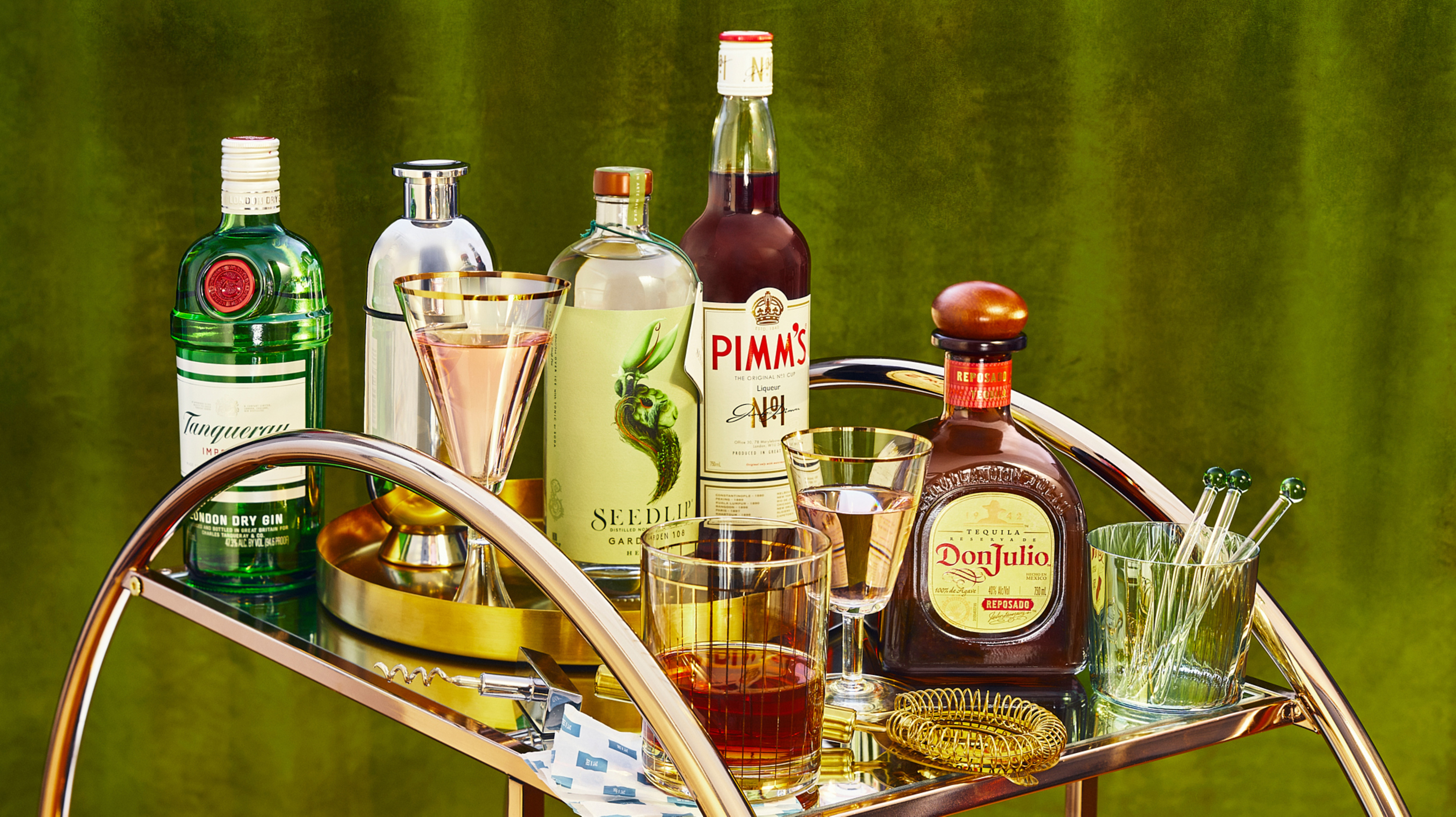 Elegant bar cart showcases gin, tequila, and other liquor bottles alongside glassware and a gold cocktail strainer against a green backdrop.