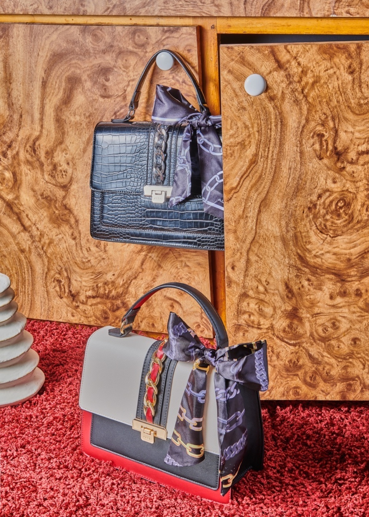 Two stylish handbags displayed against a wooden cabinet and red carpet. One black and one white and red, both adorned with scarves.