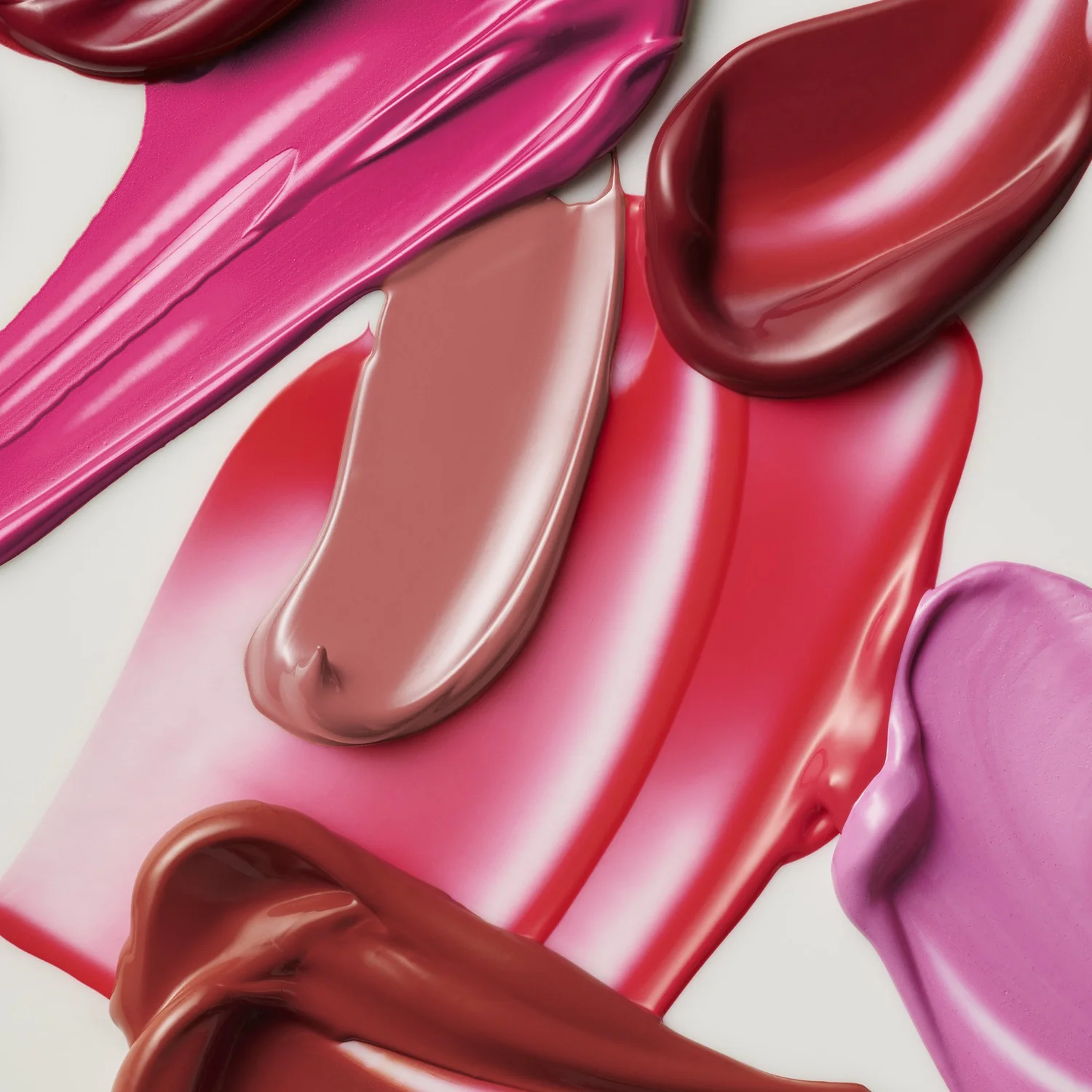Close-up of smears of pink, red, and mauve lipsticks and lip glosses on a white surface, showcasing textures and rich colors.