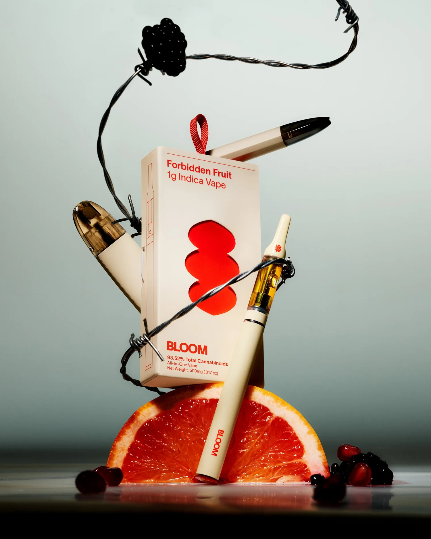 Still life of a Forbidden Fruit Indica Vape cartridge and packaging, featuring an orange slice, berries, and barbed wire.