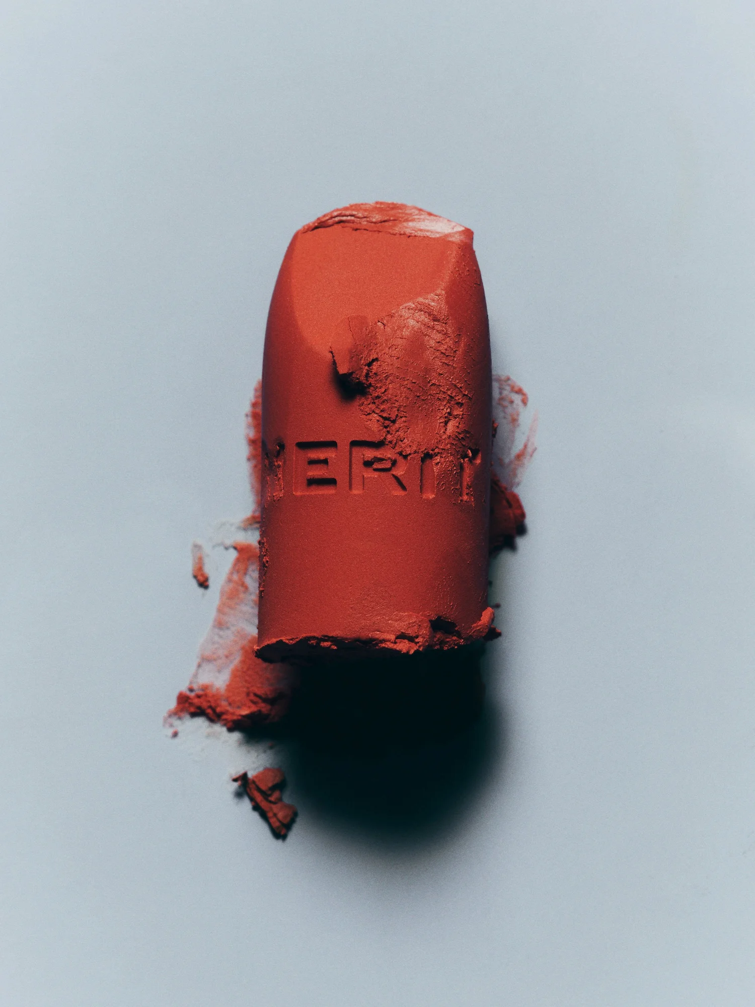 Close-up of coral-red lipstick bullet with the brand name 'Merit' embossed on the side, resting against a pale blue background.