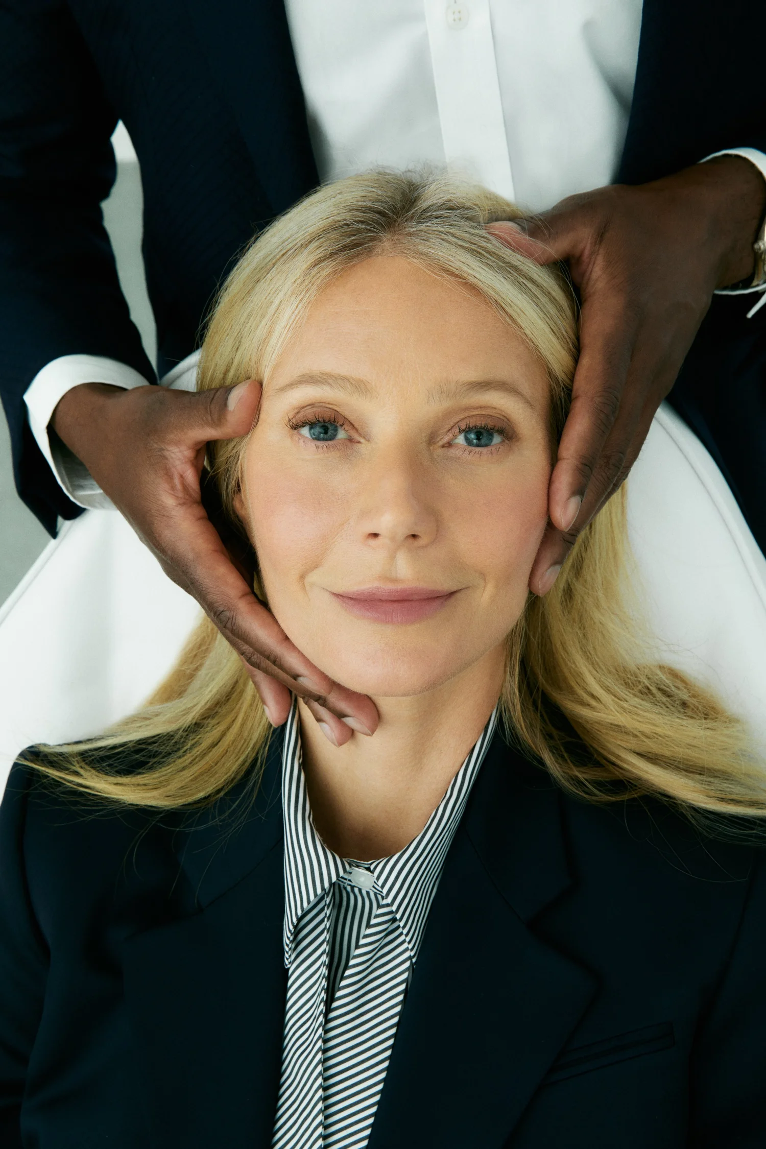 Close-up portrait of a blonde woman in a blazer having her face touched by a person in a dark suit, shot in a bright photo studio.
