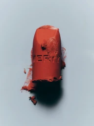 A piece of textured terracotta matte lipstick embossed with lettering standing upright on a pale grey-blue background.