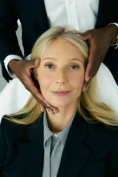 Gwyneth Paltrow seated wearing a dark blazer and striped shirt with hands framing her face.