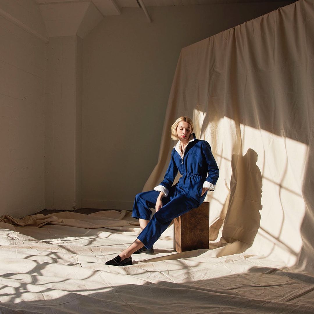 Adult female seated indoors wearing a blue jumpsuit and black loafers on a canvas backdrop with shadows from a light source.