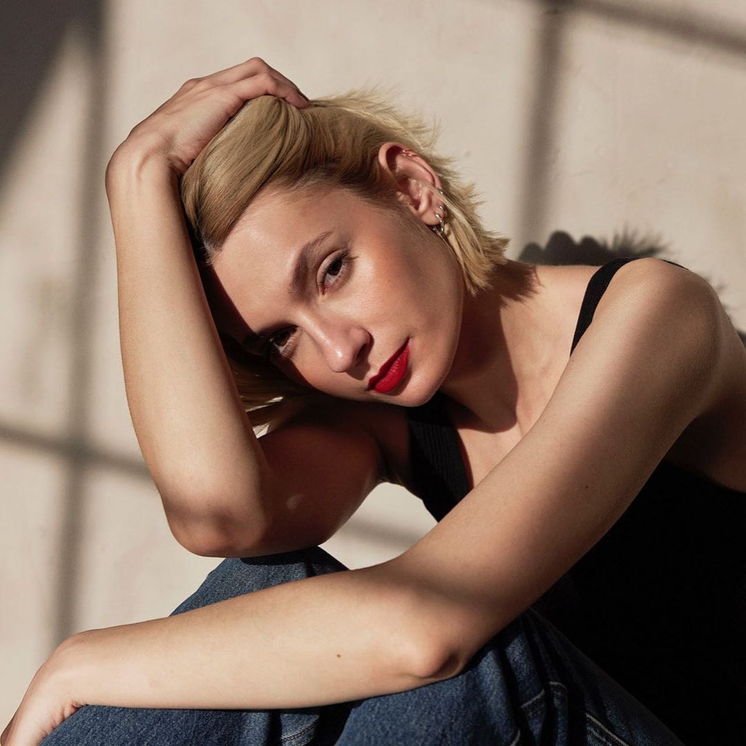 Blonde woman with red lipstick poses with a hand on her head in front of a neutral wall with window shadow patterns.