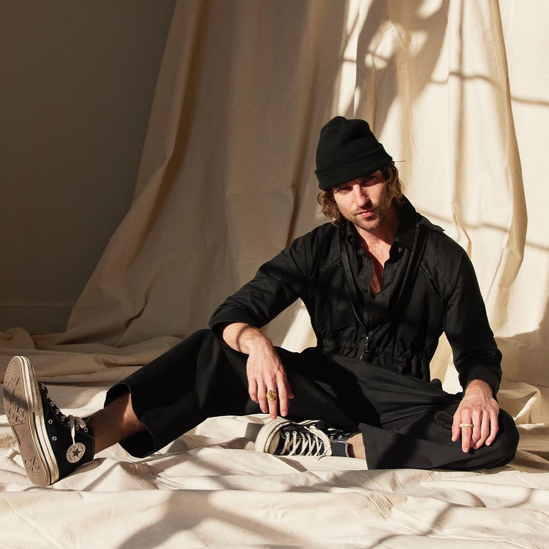 Male model in black outfit and beanie poses sitting in front of a cream-colored backdrop inside a photo studio with soft, diffused lighting.