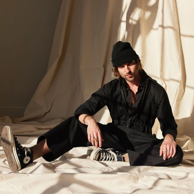 Male model in black outfit and beanie poses sitting in front of a cream-colored backdrop inside a photo studio with soft, diffused lighting.