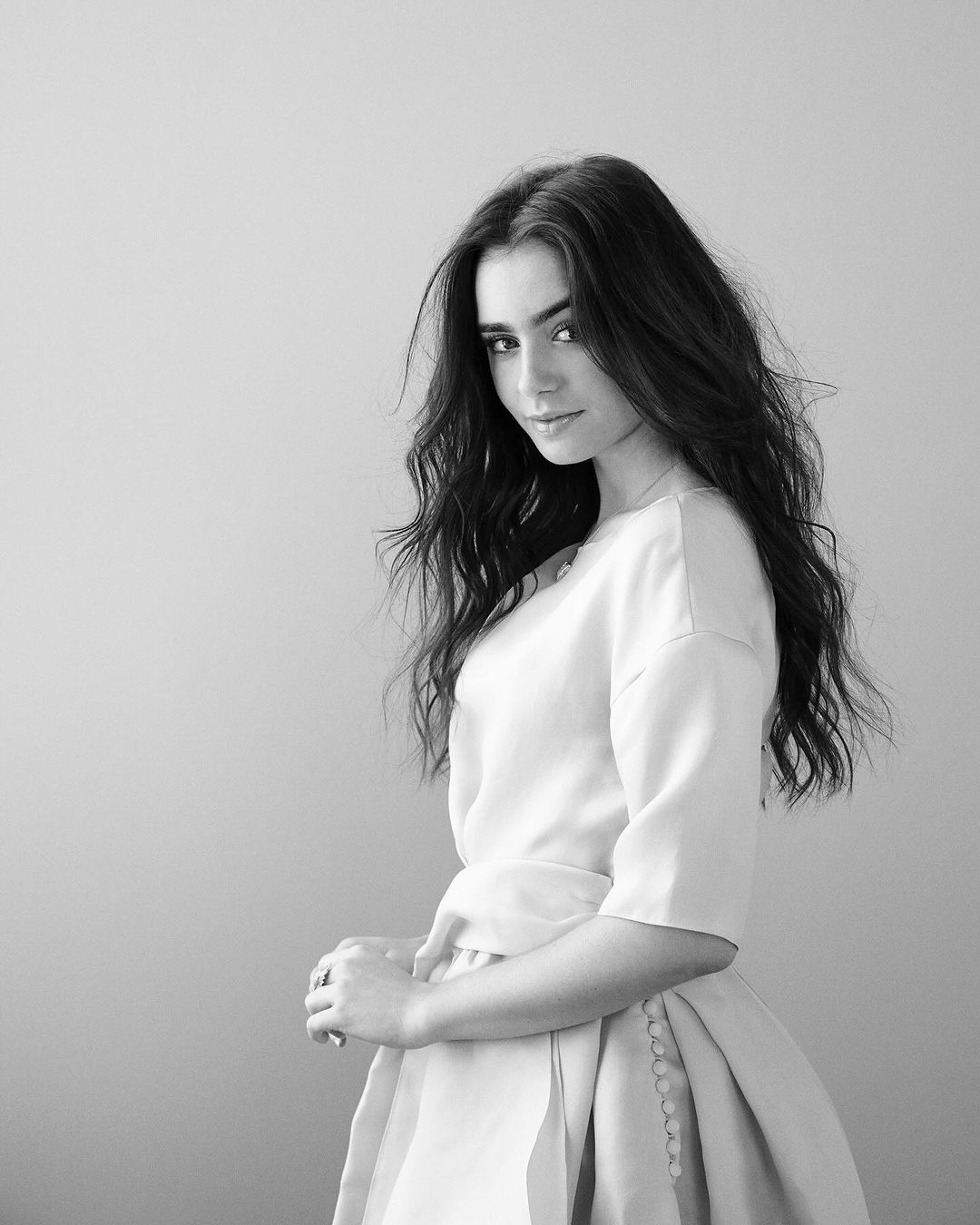 Black-and-white portrait of a woman with long, dark wavy hair, wearing a light-colored top and skirt, standing against a plain background.