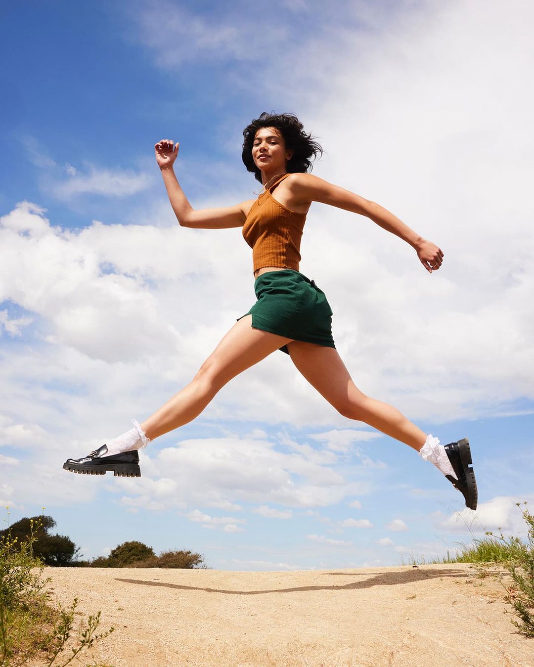 A young adult female with curly hair mid-leap against a blue sky with clouds, wearing a ribbed top, skirt, socks, and black loafers.