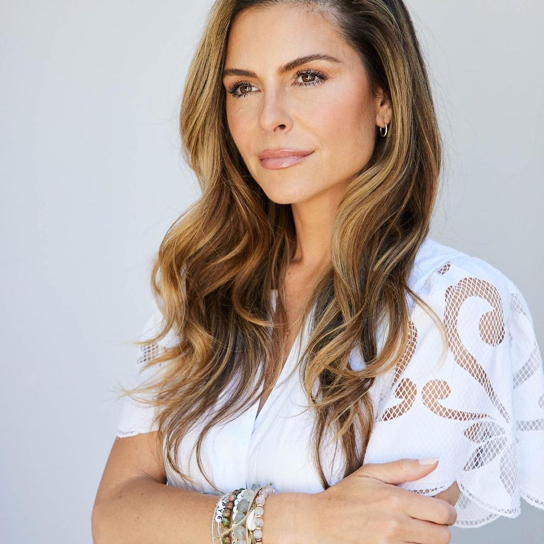 Portrait of an adult woman with long brown hair, wearing a white blouse and bracelets, posing against a neutral background in soft light.