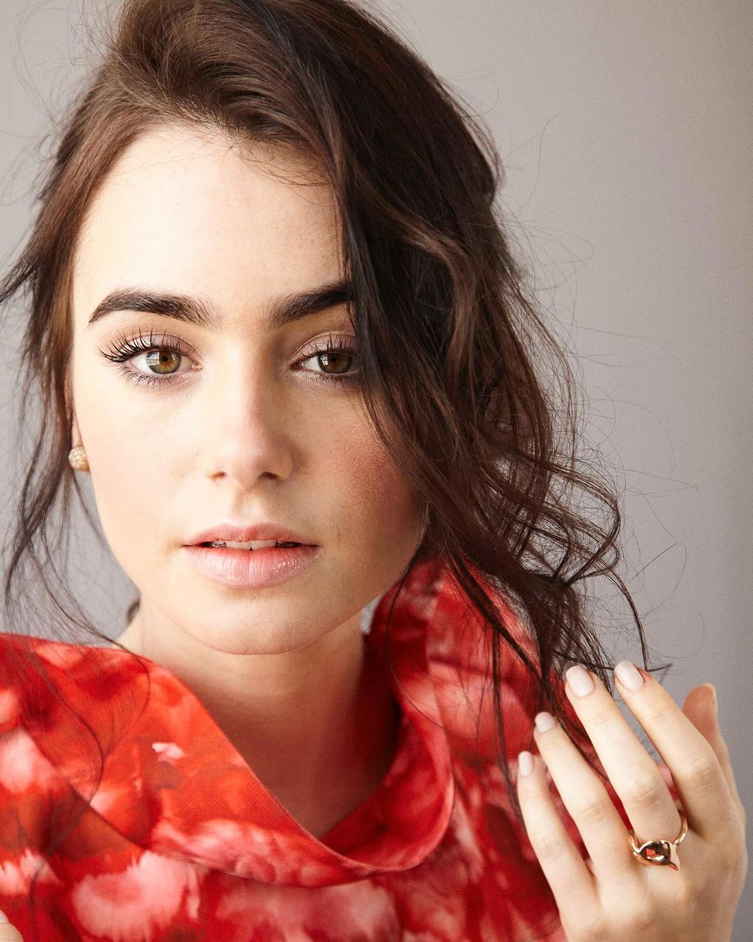 Portrait of a woman with dark hair and light eyes wearing a red and white floral top, posing in front of a neutral background.