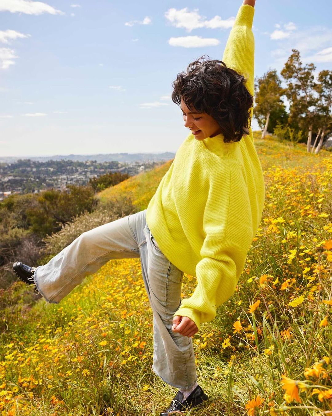 A young woman in a yellow sweater and jeans stands in a field of wildflowers, smiling against a background of hills and sky.