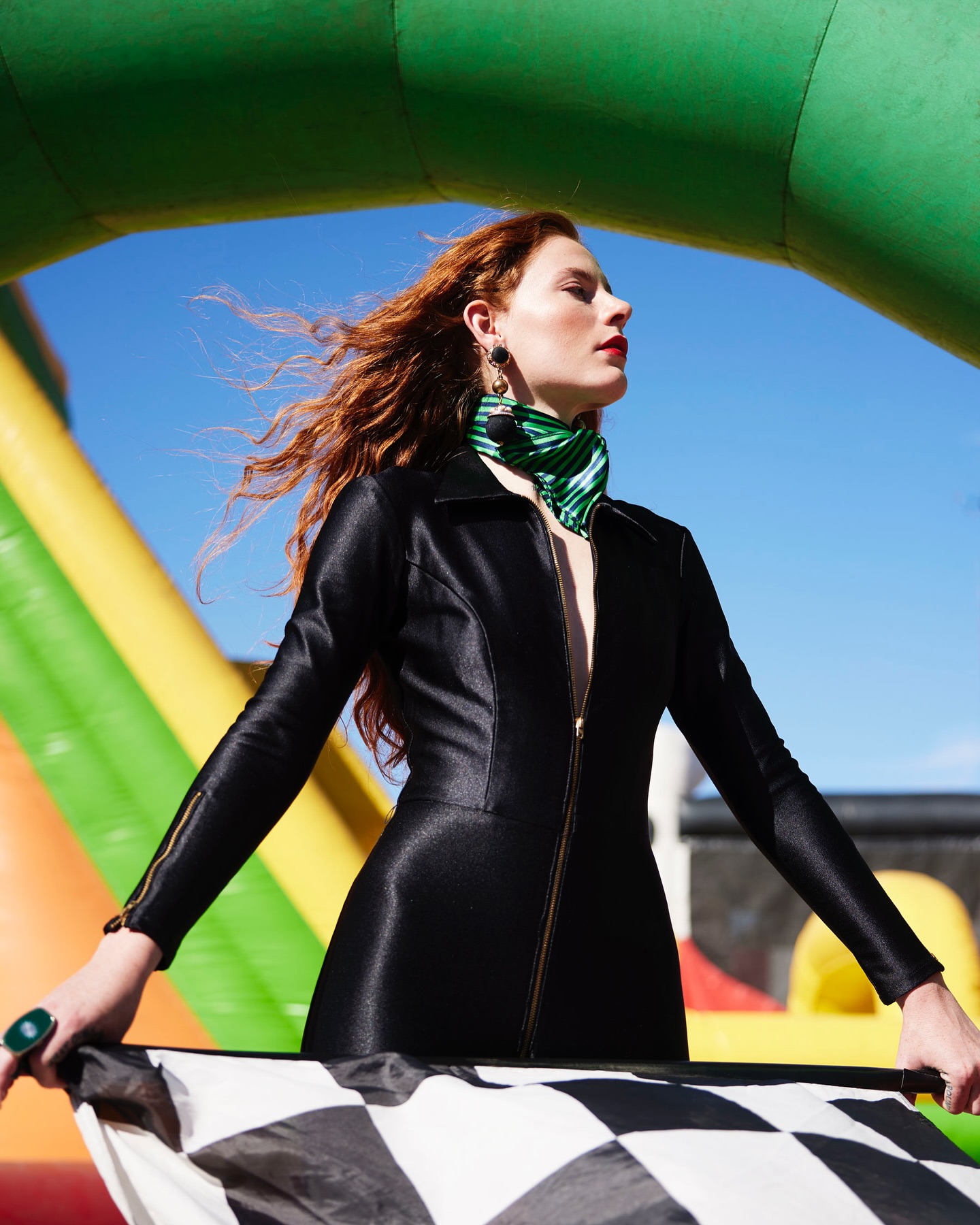 A red-haired woman in a black jacket holds a checkered flag, posing in front of an inflatable structure against a blue sky.