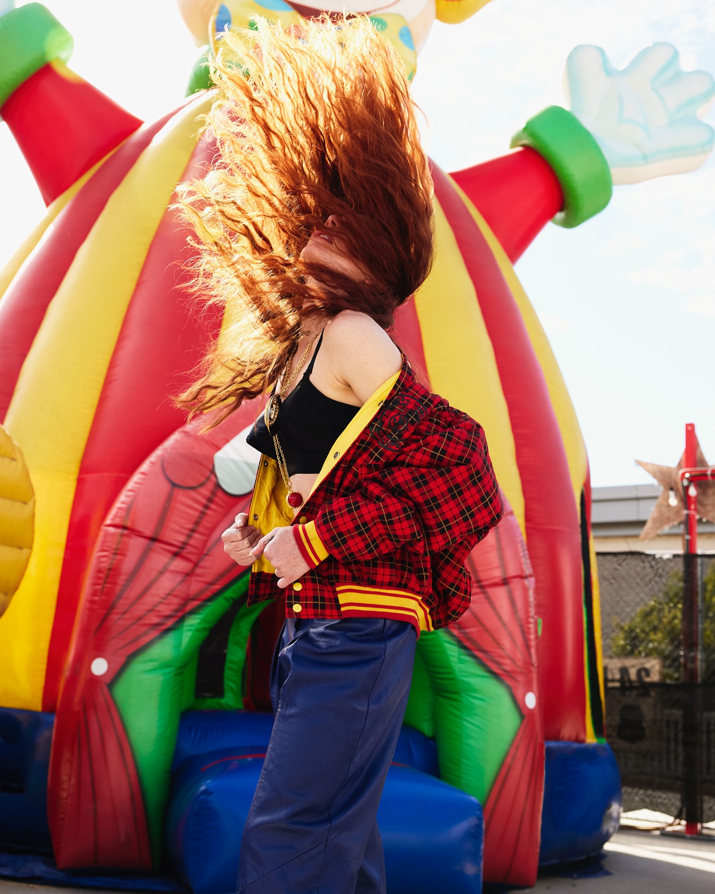 Woman with red hair poses outdoors in front of a colorful inflatable clown. She wears a plaid jacket and blue pants in full sunlight.