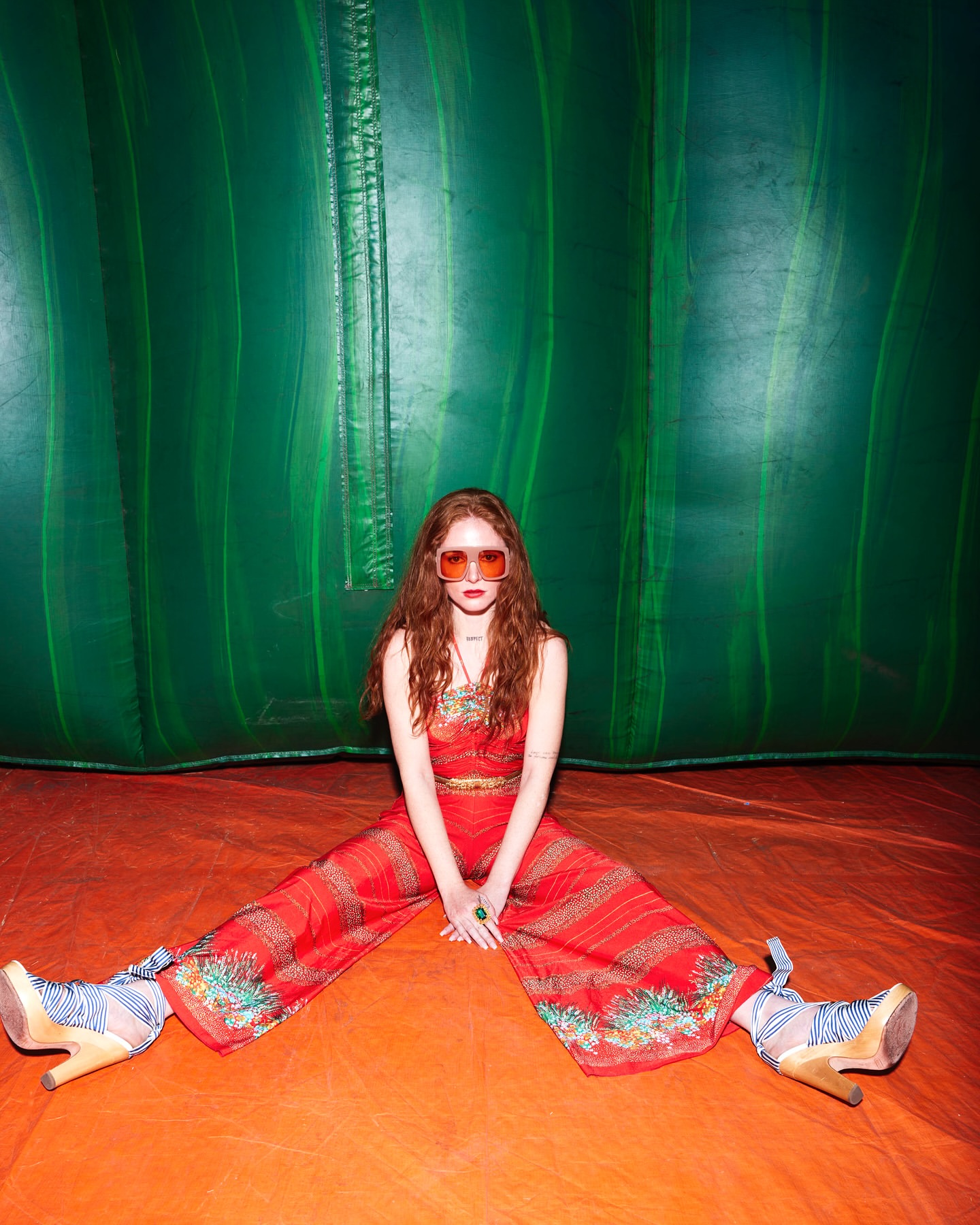 A red-haired woman in a red jumpsuit and oversized sunglasses sits cross-legged on an orange surface against a green background.