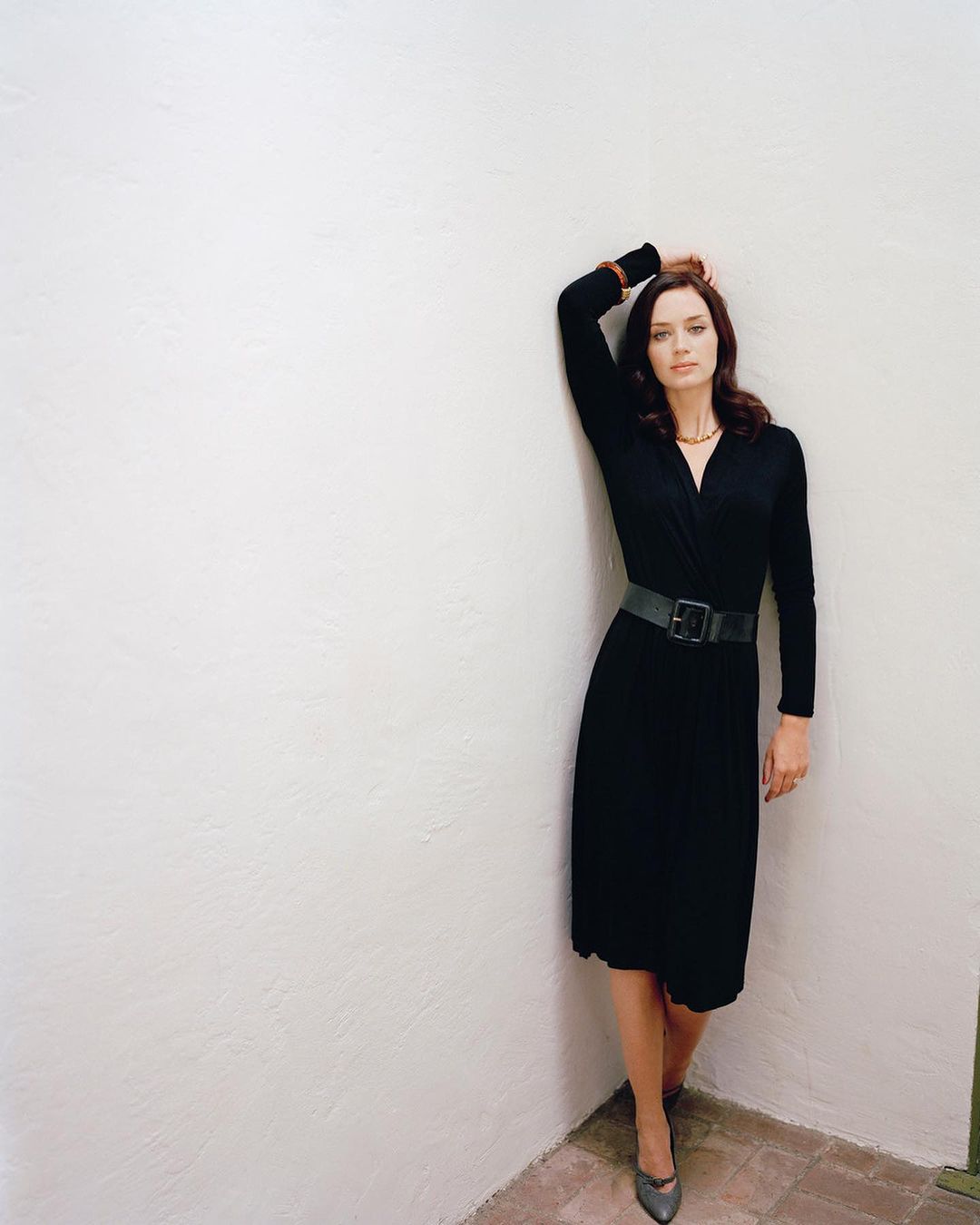 Elegant woman in a black dress poses against a white wall, hand raised to her head, standing indoors with sophisticated style and grace.