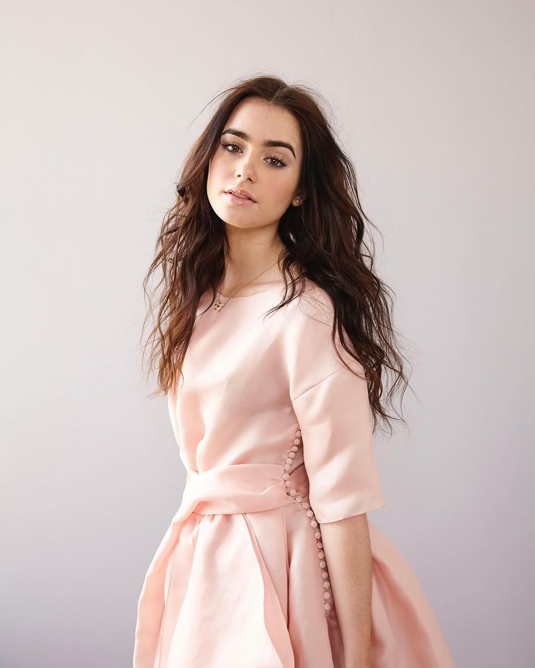 Portrait of a brunette woman in a pastel pink dress with an A-line skirt, posing indoors in front of a neutral background.