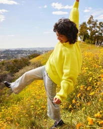Smiling person wearing a bright yellow sweater and light blue jeans posing in a sunlit meadow of yellow wildflowers