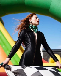 Red-haired woman in a black zipped jumpsuit holding a checkered flag outdoors against an inflatable structure.
