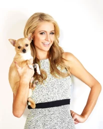 Paris Hilton holding a small tan Chihuahua while wearing a patterned sleeveless dress against a plain white background.
