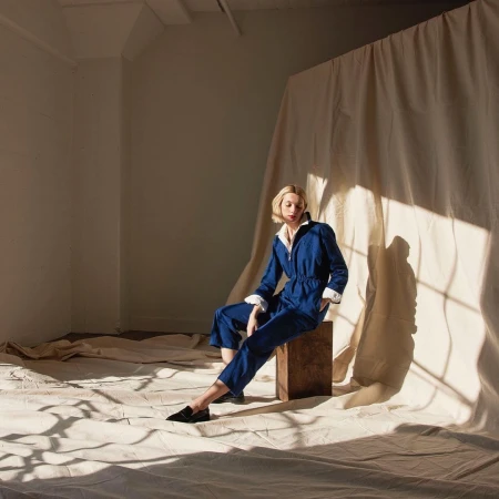 Blonde woman wearing a blue jumpsuit seated on a wooden block against a draped fabric backdrop with diagonal window light shadows