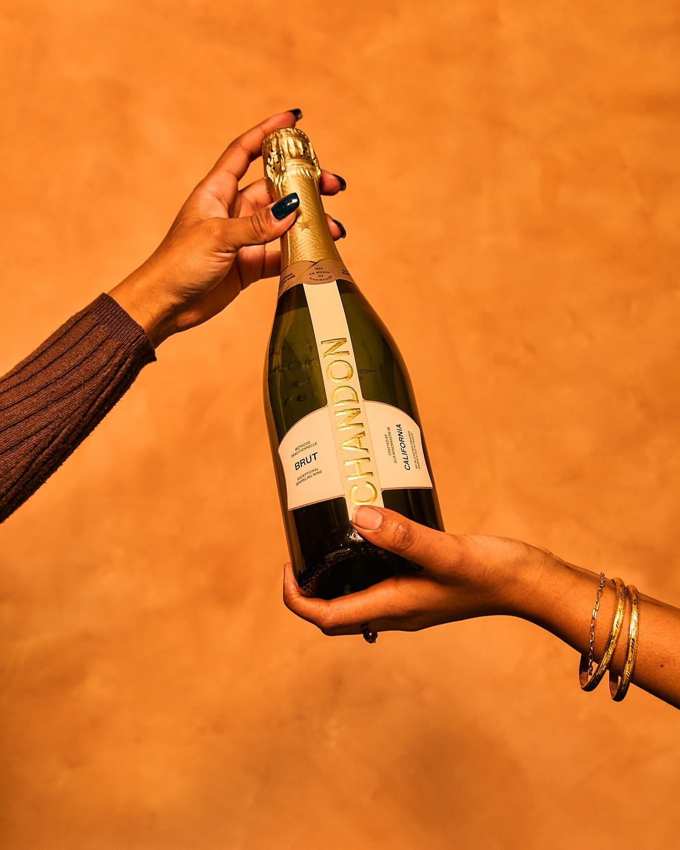 Two dark-skinned hands holding a bottle of Chandon sparkling wine against a textured orange background. The bottle's label is prominently displayed.