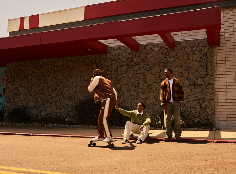 Three adult men outside of a building; one man is skateboarding while another is holding his hand and the third watches.