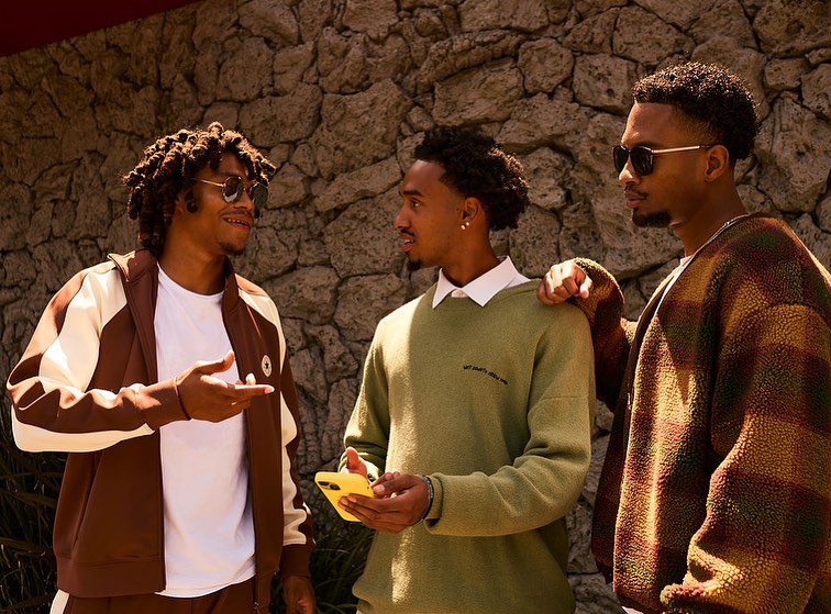 Three young Black men in casual sweaters and jackets stand in an outdoor setting with a stone wall, two wearing sunglasses and holding a phone.