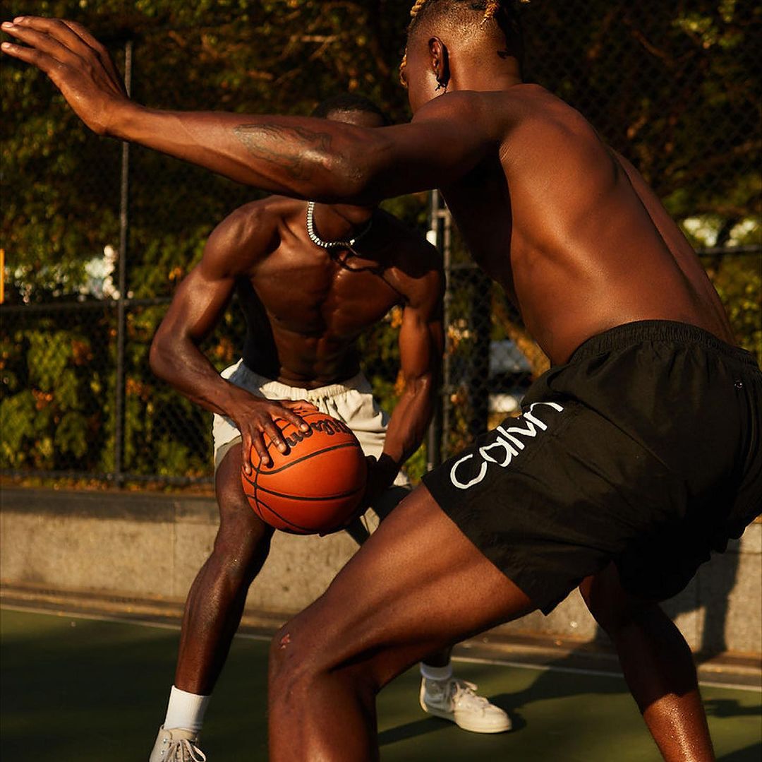 Two athletic, bare-chested men playing basketball on an outdoor court on a sunny day, with one dribbling the ball. 