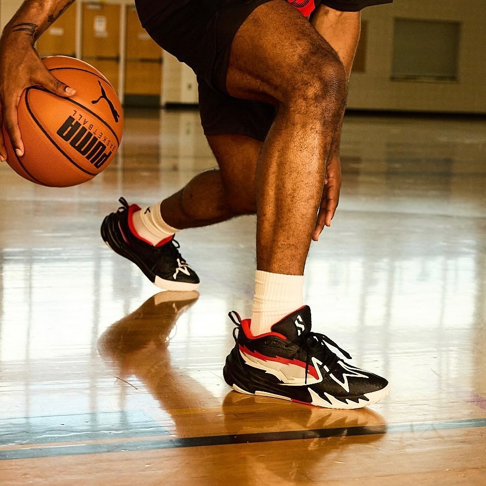 African American man dribbles Puma basketball in gymnasium, wearing black and white athletic shoes, white socks, and black shorts.